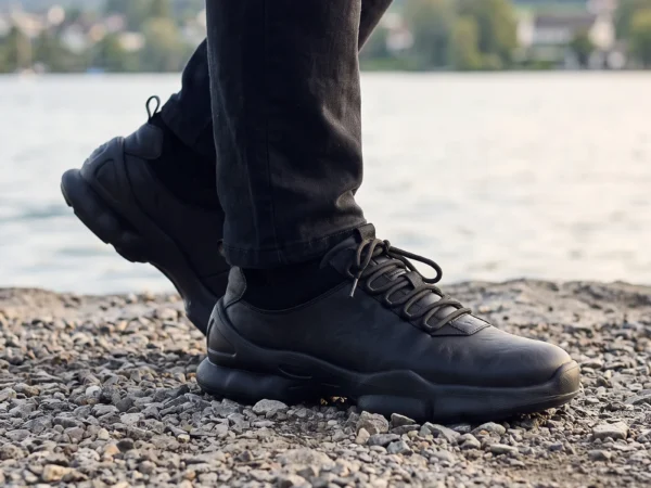 leather breathable hiking walking shoes lightweight non-slip outdoor sneakers in black, worn on foot on a lakeside shore, soft daylight lifestyle photo