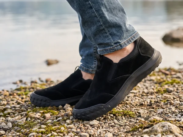 slip-on work safety shoes lightweight suede anti-slip driving shoes in black, worn on foot on a lakeside shore, soft daylight lifestyle photo